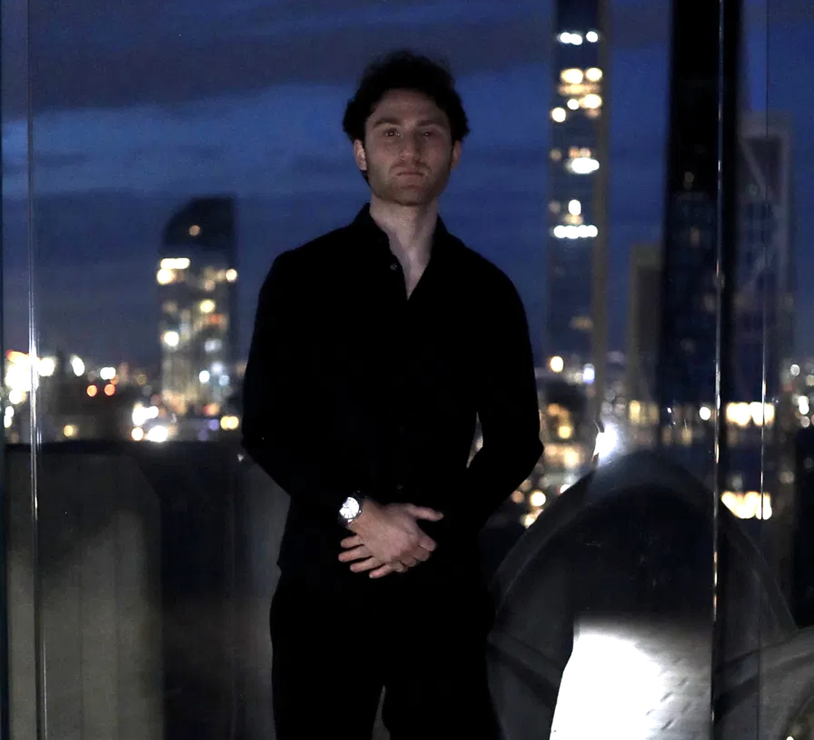 Matthew Hannani standing at a window above a city skyline at night